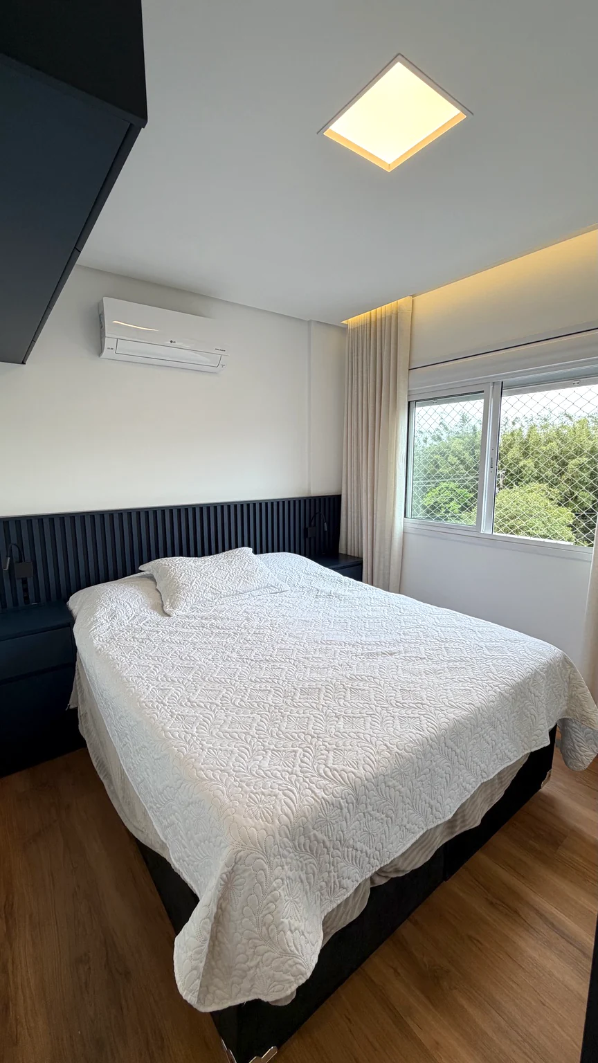 Quarto de casal, painel de cabeceira, janela e aparelho de ar-condicionado.
